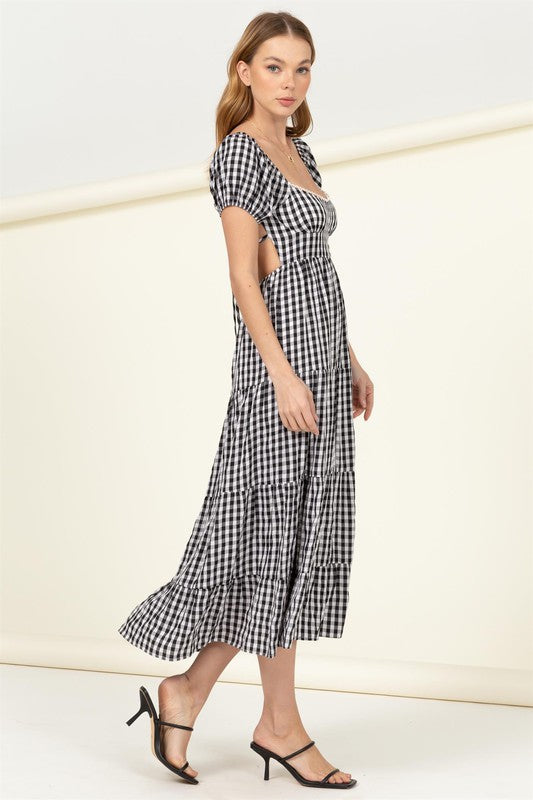 Wearing the "Somewhere to Go Tie-Back Gingham Print Maxi Dress," which features a black and white gingham print with puffed sleeves, a tiered skirt, and elegant tie-back detail, a person poses stylishly paired with black sandals.