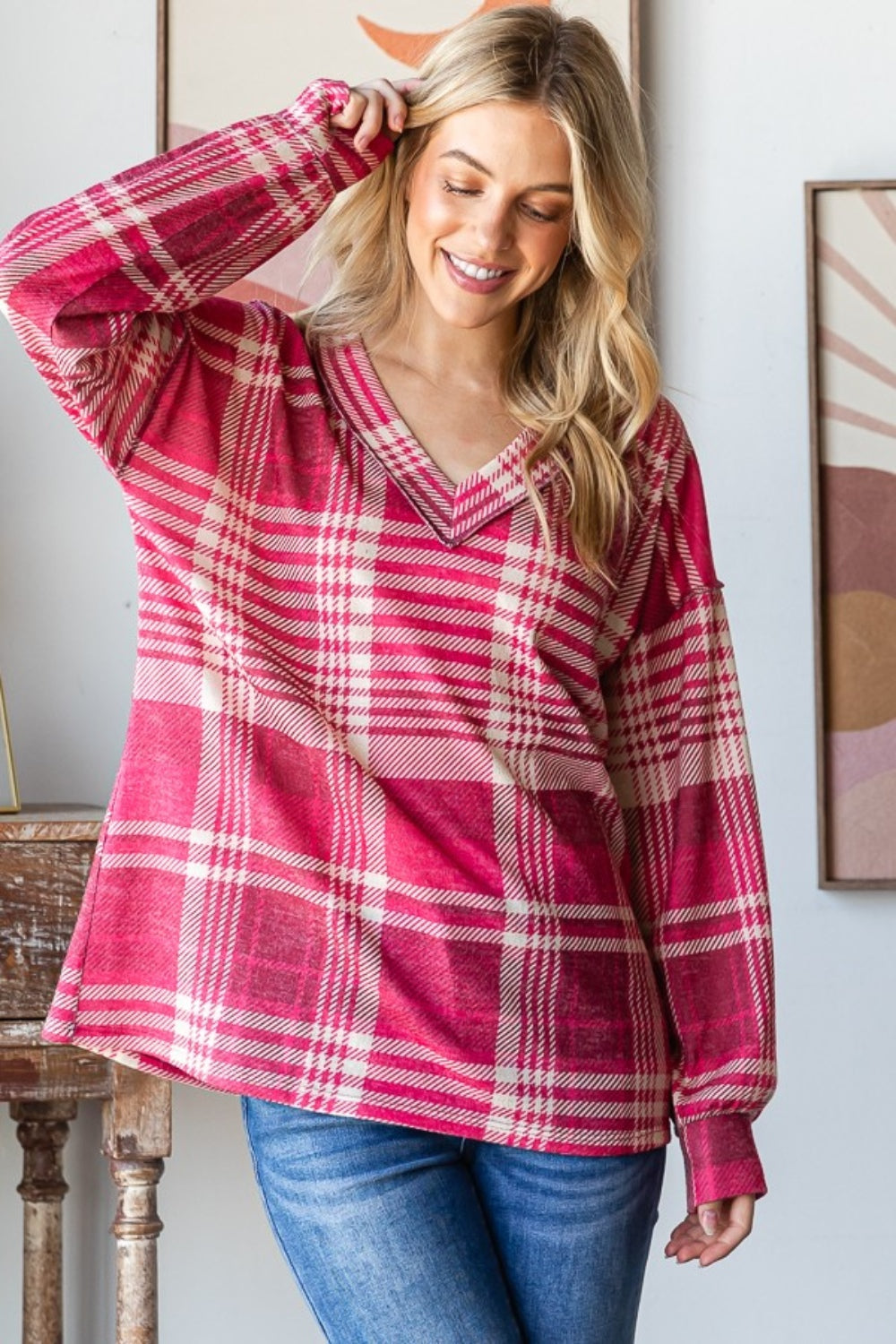 A person with long blond hair smiles while wearing the Heimish Plaid Drop Shoulder V-Neck T-Shirt in red and white and a pair of blue jeans, leaning against a wooden table with framed pictures in the background.