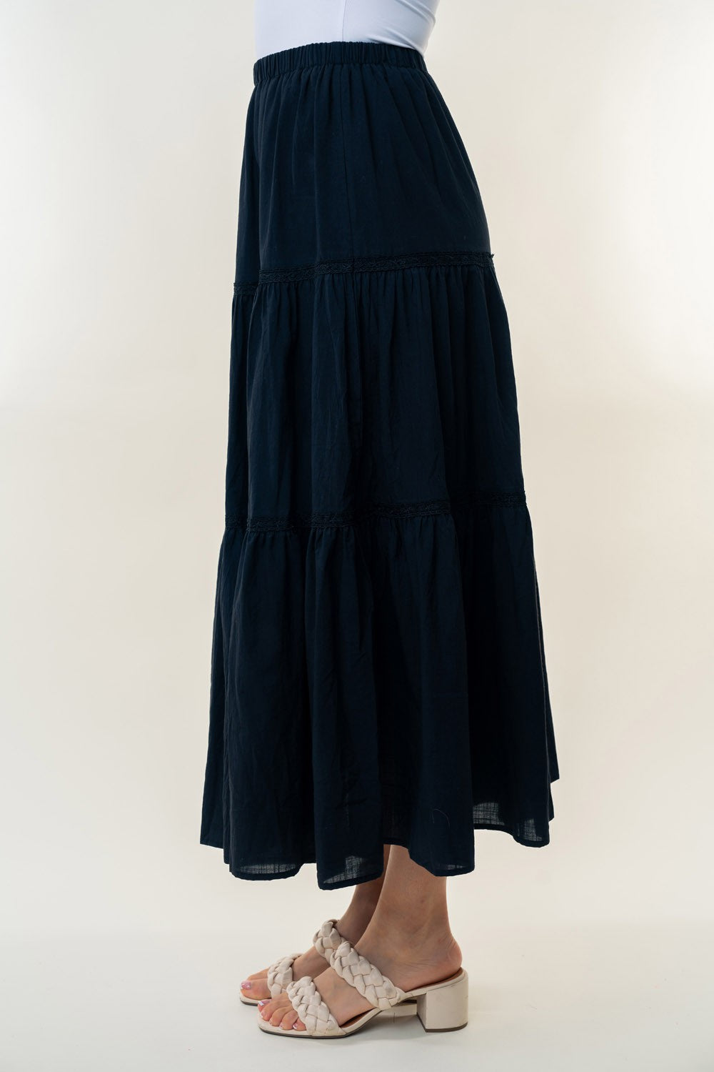 A long, White Birch High Waisted Tiered Maxi Skirt in navy blue, offering a versatile piece perfect for a bohemian look. It's worn by a person paired with white heeled sandals. The background is plain and light-colored.