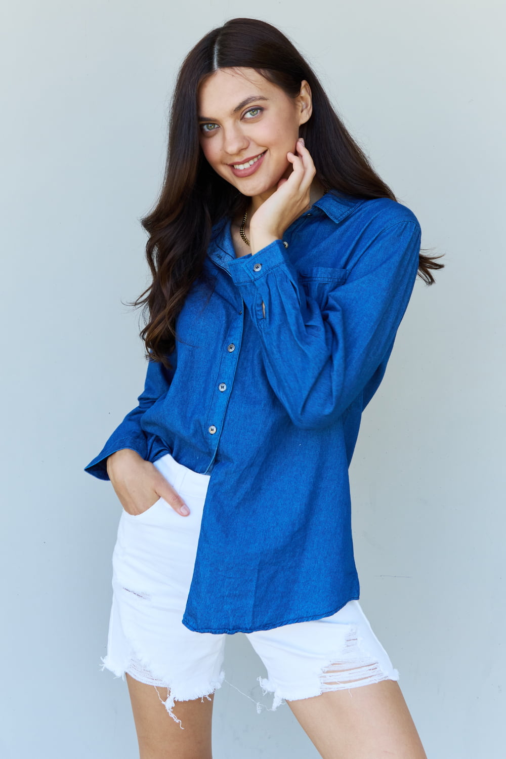 A person standing against a plain background, wearing the Doublju Blue Jean Baby Denim Button Down Shirt Top in Dark Blue and white shorts, smiling and holding a hand to their hair.