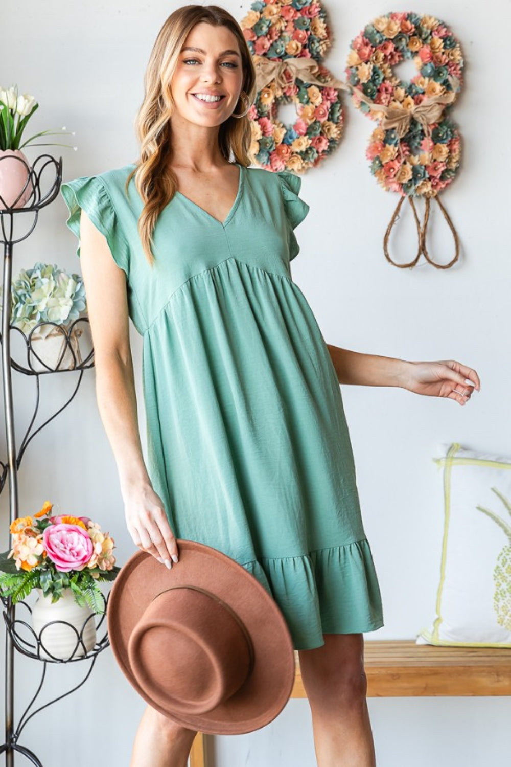 A person in a Heimish Full Size Short Sleeve V Neck Ruffled Hem Dress stands indoors, elegantly holding a hat. The floral decor in the background complements the dress's feminine charm.