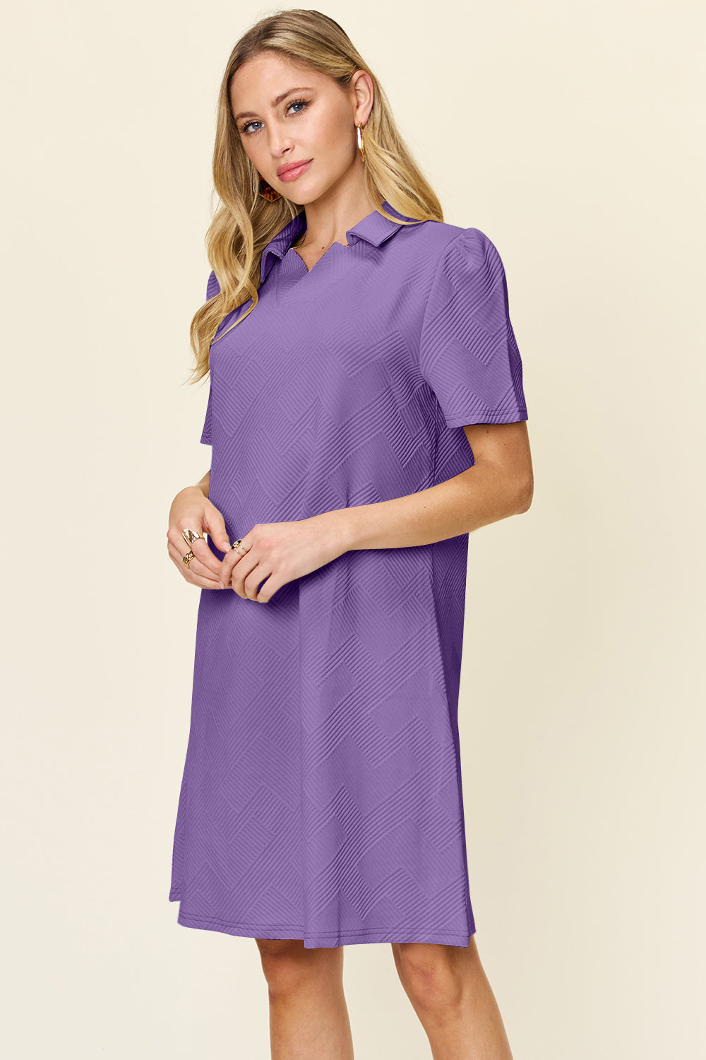 A woman wearing the Double Take Full Size Texture Collared Neck Short Sleeve Dress, featuring a geometric pattern in purple and made from soft polyester, stands against a neutral background. She has long, light hair and is looking slightly to the side. The dress is available in sizes S-3XL and is easy to care for—just machine wash cold.