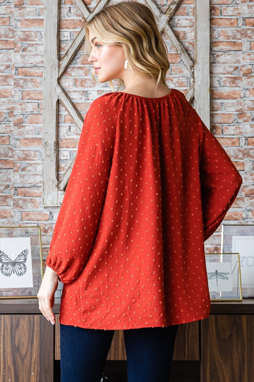 A woman wearing a Heimish Full Size Swiss Dot Off Shoulder Top paired with dark pants poses indoors against a brick wall decorated with butterfly artwork.