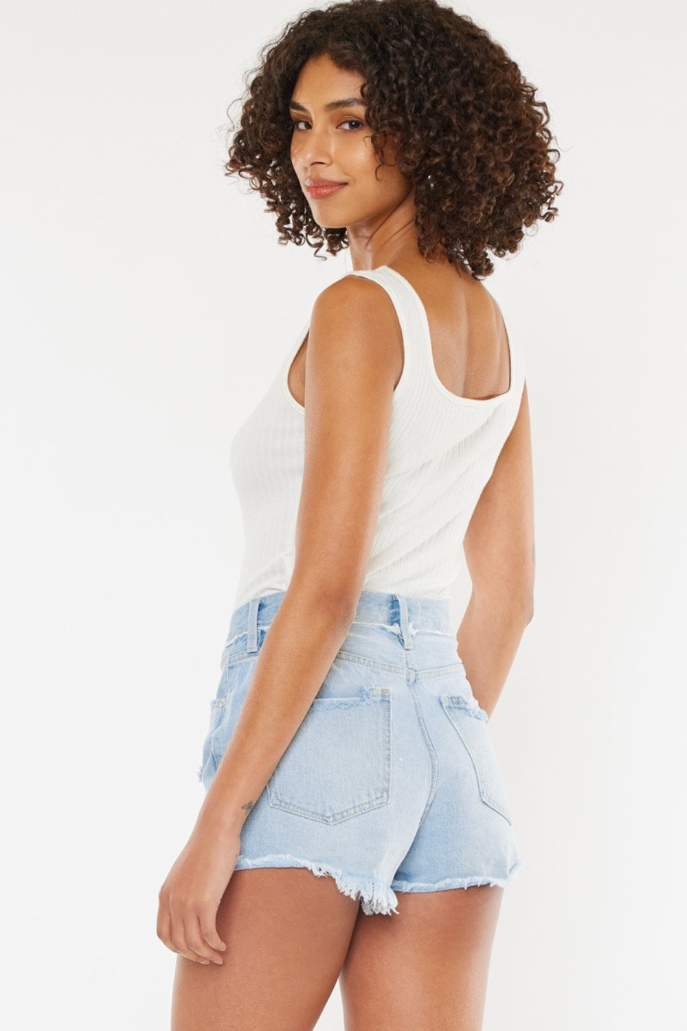 A person wearing a white tank top and Kancan Distressed Button Fly Denim Shorts poses against a plain background, showcasing a quintessential summer wardrobe staple.