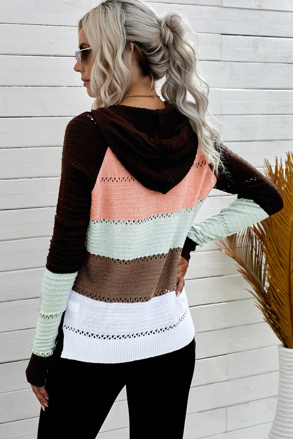 A person wearing the Brown Zipped Front Colorblock Hollow-out Knit Hoodie, featuring a multi-colored striped design with alternating bands of brown, pink, white, and gray. Seen from the back, the sweater showcases the hood and finely knitted texture. The person has long wavy hair—a perfect look for a bonfire on the beach.