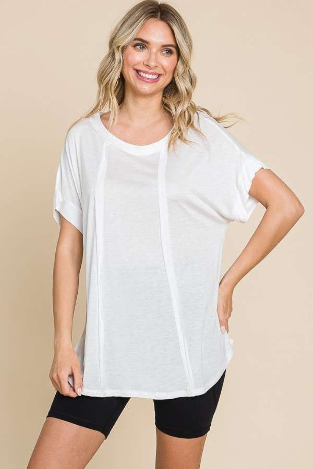 Woman in a white Culture Code Round Neck Short Sleeve T-Shirt and black shorts, posing against a beige background with one hand on her hip. This versatile staple makes everyday wear effortlessly chic.