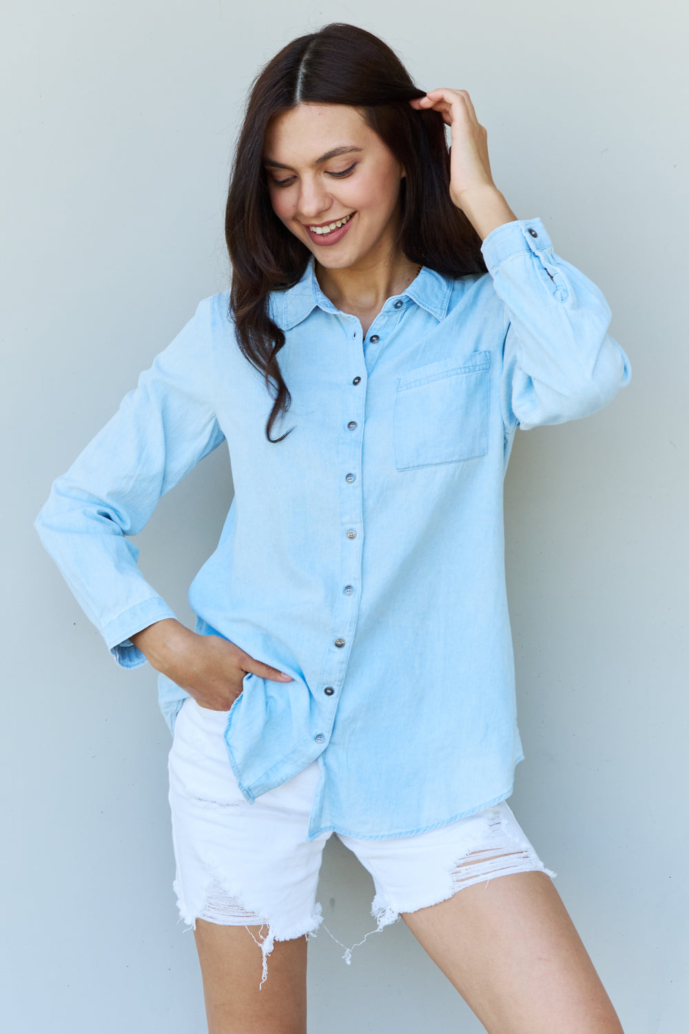 A woman wearing the Doublju Blue Jean Baby Denim Button Down Shirt Top in Light Blue, paired with white ripped shorts, strikes a pose with one hand in her pocket against a plain background, highlighting the classic button-up silhouette.