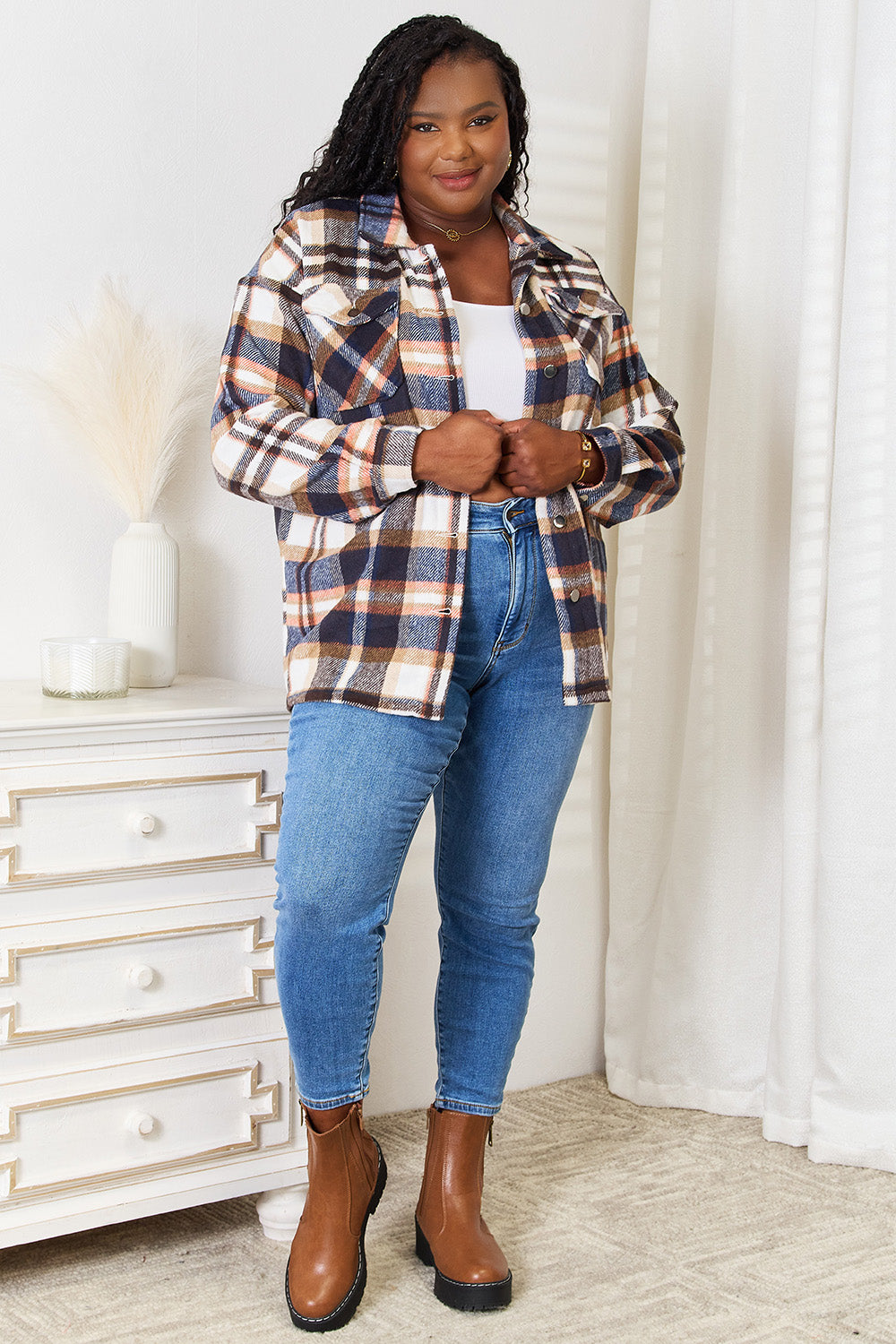 A person with long braided hair wearing the Double Take Plaid Button Front Shirt Jacket with Breast Pockets over a white top and blue jeans stands with a hand in their pocket, posing in an indoor setting. The layering of the jacket adds an effortless touch to the outfit.