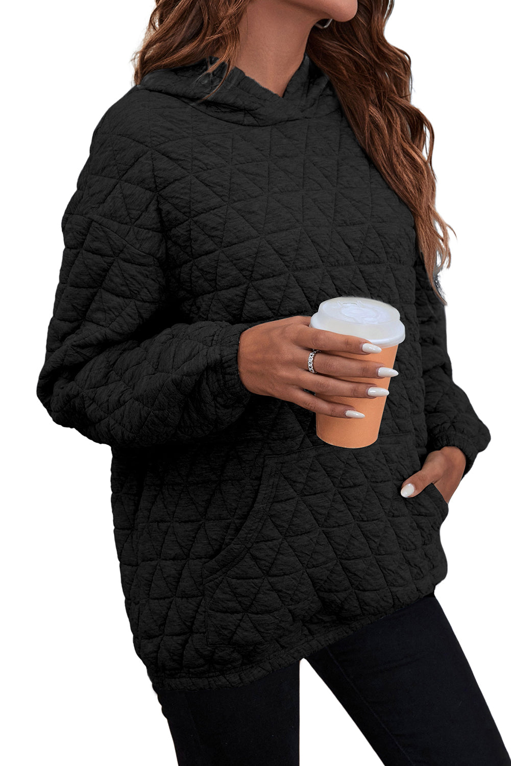 A person with wavy brown hair is standing indoors, facing a window, and wearing a Black Solid Color Quilted Kangaroo Pocket Hoodie with a drop shoulder design.