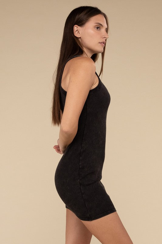 A woman is seated, wearing the Acid Washed Ribbed Mini Dress in a faded red hue, along with a necklace. Her right arm rests on a white surface as her straight hair frames her neutral expression.