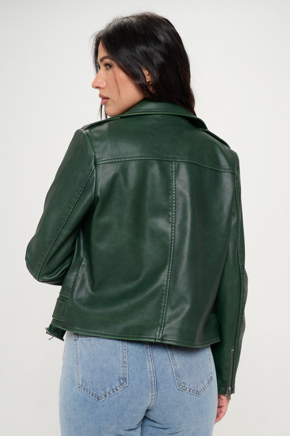 A person with long dark hair wearing a green Coalition LA Zip Up Biker Jacket with Belt and blue jeans stands against a plain background.