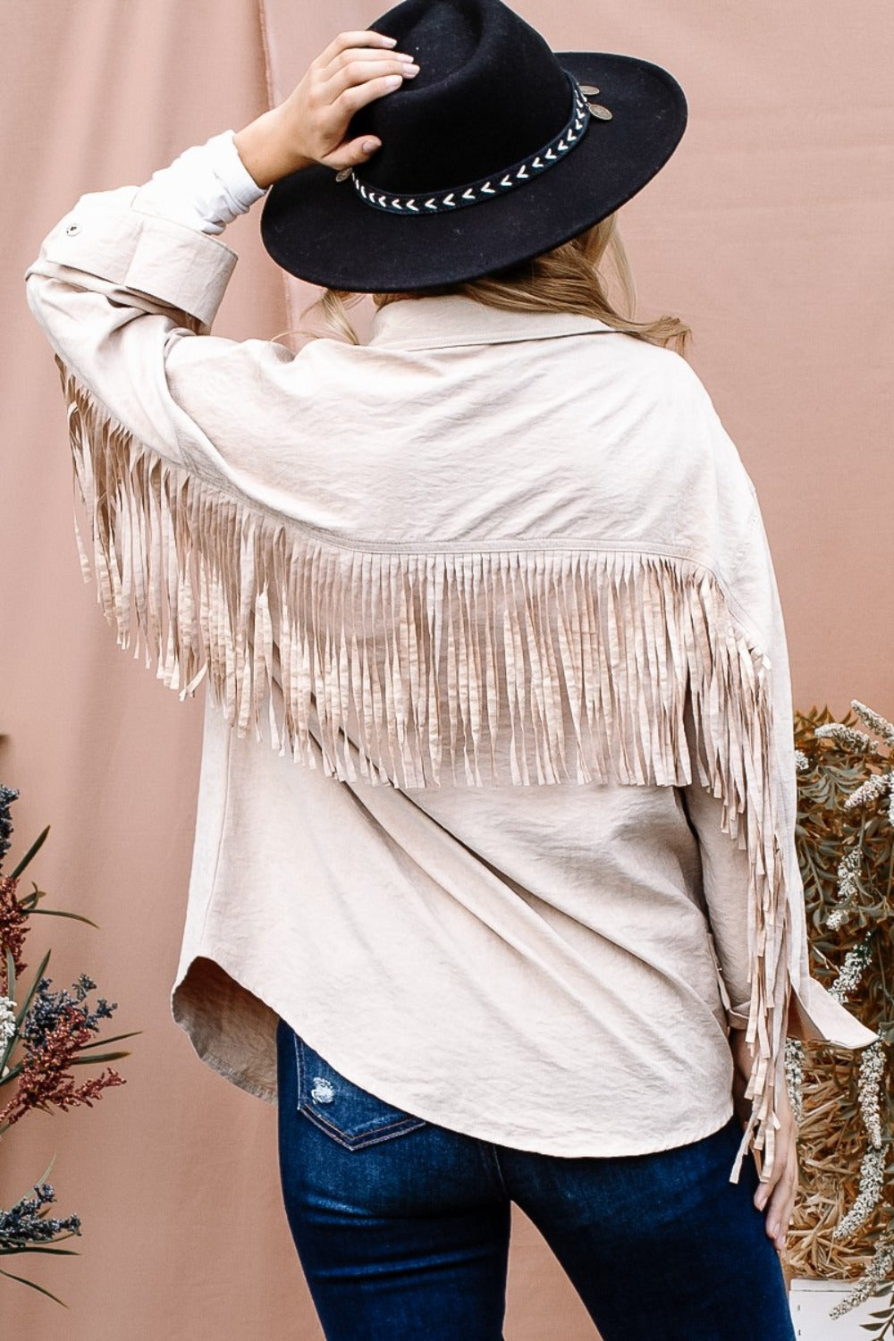 A person wearing the And The Why Full Size Fringe Back Detailed Button Down Shacket in beige, paired with a white inner layer, blue jeans, and a black hat, poses with one hand on the brim of the hat against a tan background.