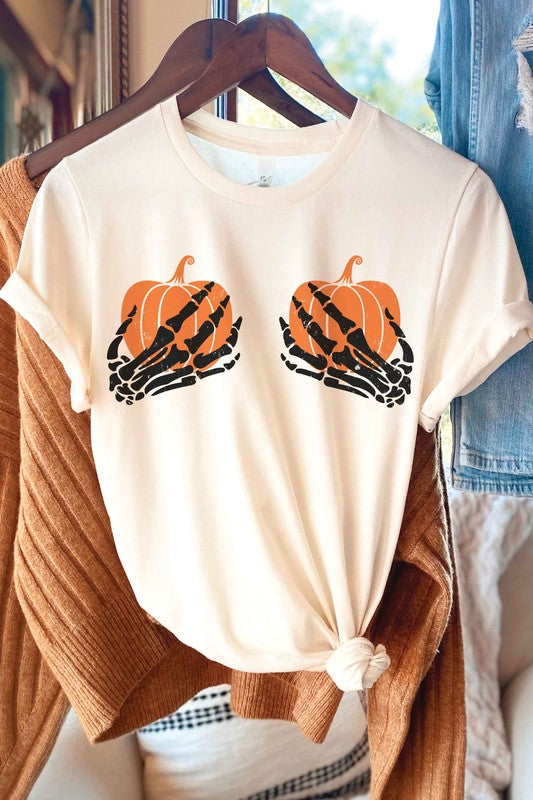 A person wearing the SKELETON HOLDING PUMPKINS Graphic Tee, which features two orange pumpkins and black skeletal hands, is sitting down. With long, wavy hair and accessorized with various bracelets and a necklace, the unisex sizing of the shirt makes it a versatile addition to any wardrobe.