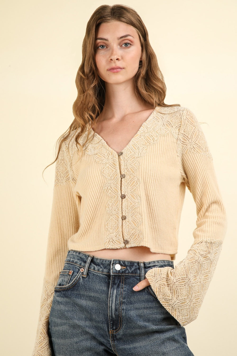 A woman with long brown hair is wearing the VERY J V-Neck Lace Detail Button Down Crop Ribbed Knit Top and blue jeans, standing against a plain background. The crop top features stylish bell sleeves that add a touch of elegance to her look.
