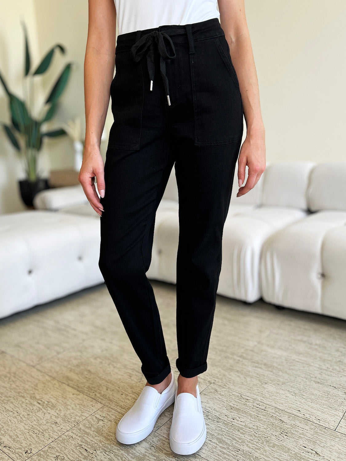 Person standing indoors, wearing a white top, black Judy Blue Full Size High Waist Double Roll Cuff Jeans, and white slip-on shoes, next to white couches and a plant in the background.