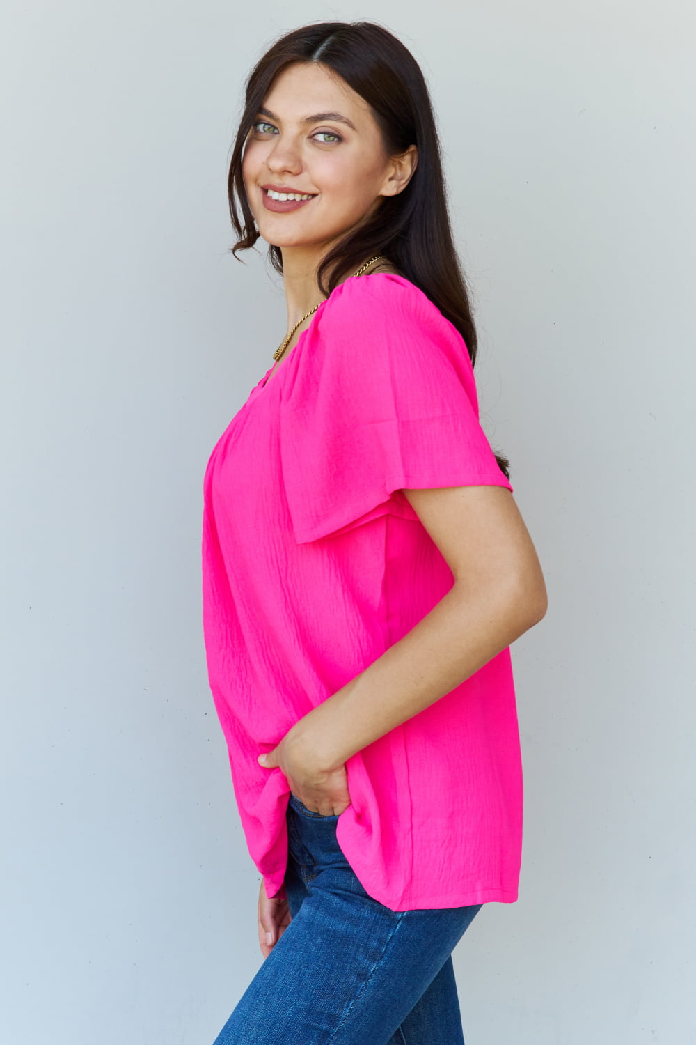 A person wearing the Ninexis Keep Me Close Square Neck Short Sleeve Blouse in Fuchsia paired with blue jeans smiles while posing against a plain background.