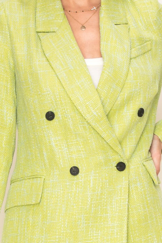 A person stands facing forward wearing the sophisticated Sugarplum Long-Sleeve Double-Breasted Blazer in green, which features a notched collared neckline, over a white top paired with matching green shorts. Their hands rest in the blazer pockets.