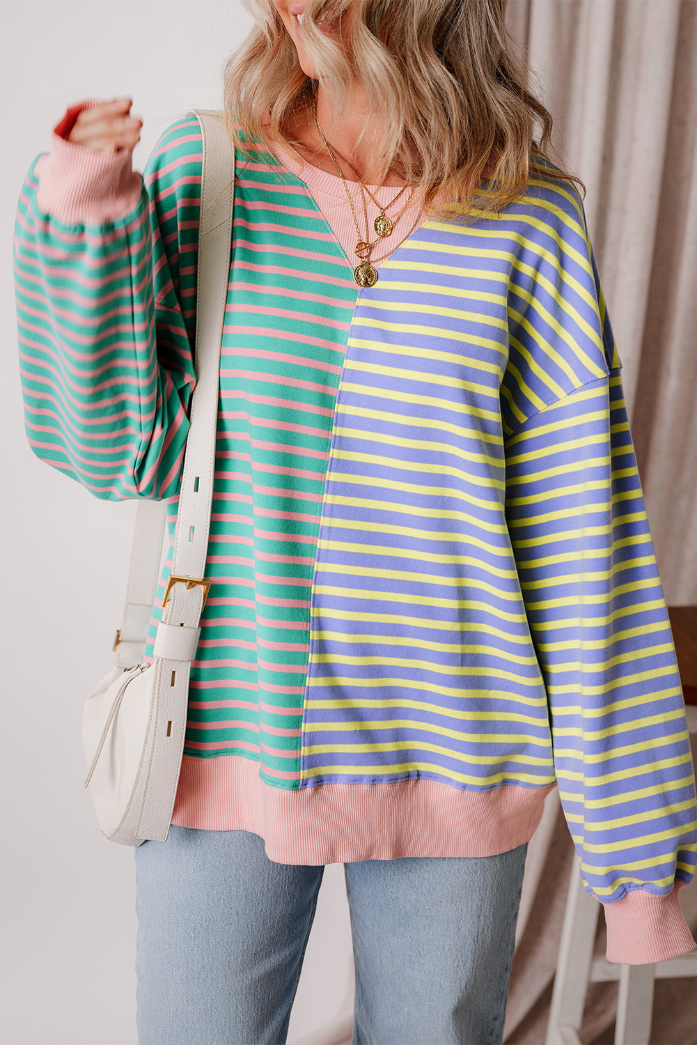 A person with wavy hair is seen from behind wearing a Casual Stripe Colorblock Drop Shoulder Oversize Sweatshirt in pastel colors and blue jeans. They are carrying a white shoulder bag, showcasing a trendy color block design.