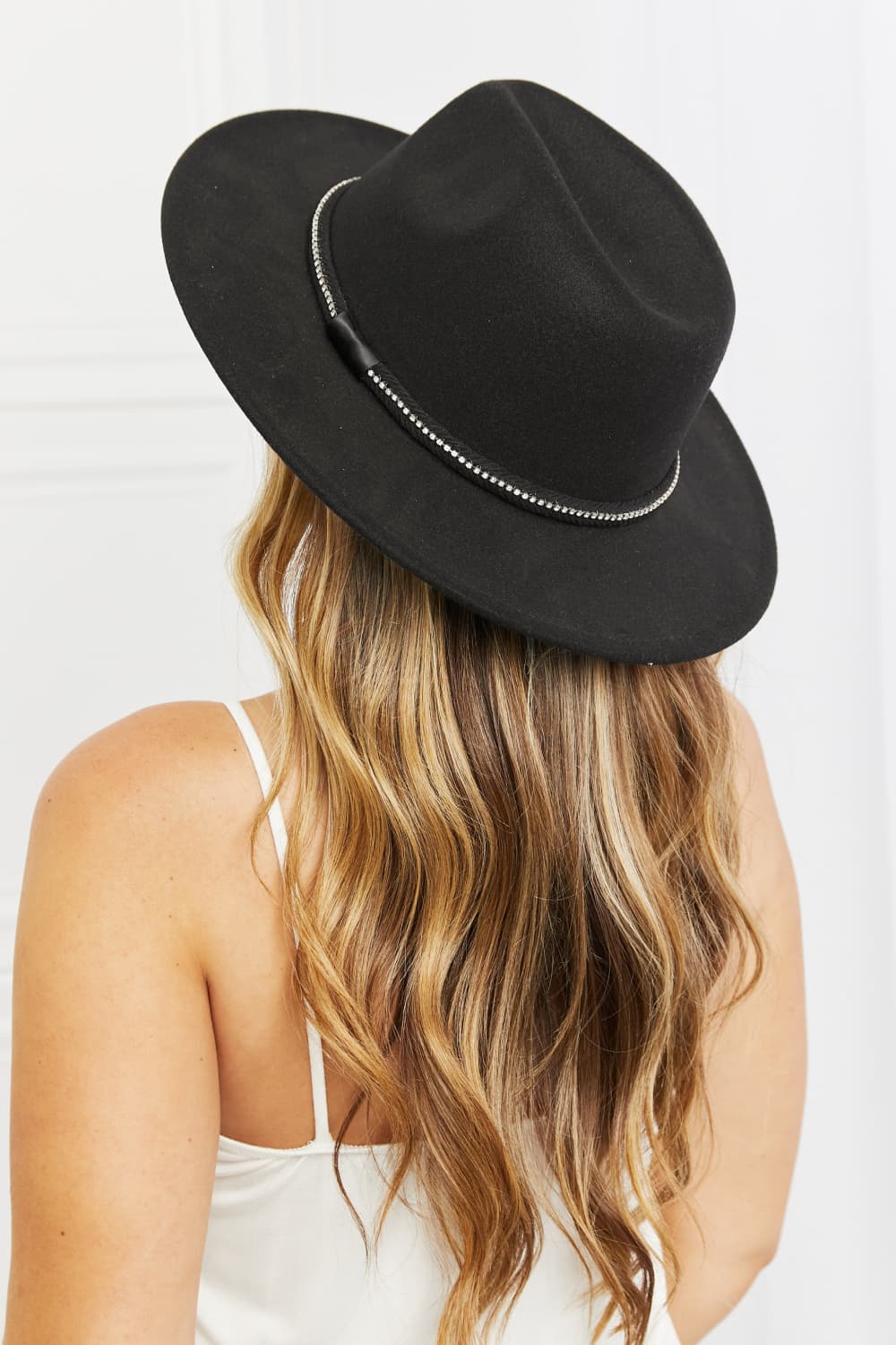 Person with long wavy hair wearing the Fame Bring It Back Fedora Hat, which features a black wide brim and rhinestone detailing, paired with a white tank top, viewed from the back.