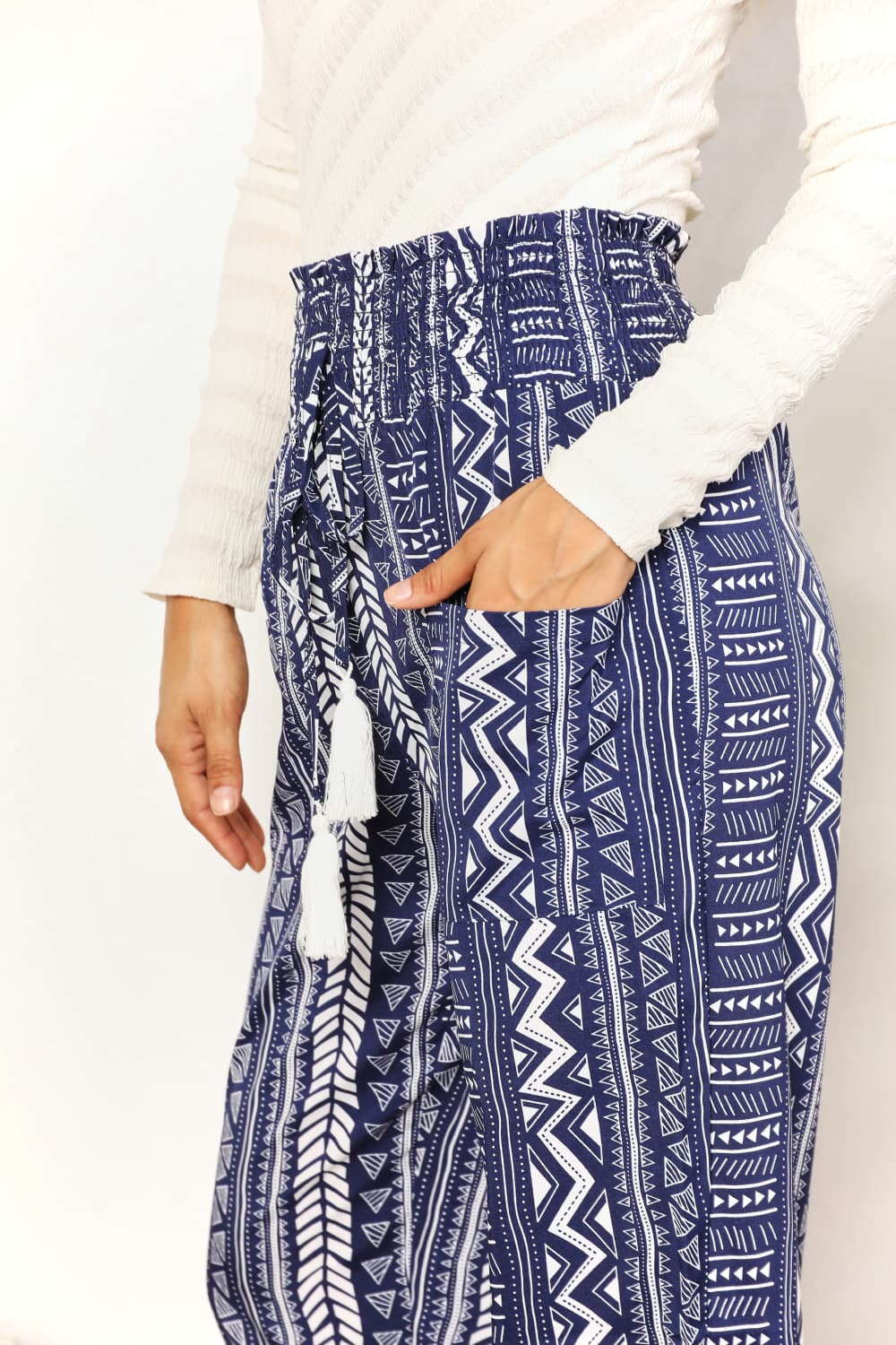 A person wearing a white long-sleeve top and Perfee Geometric Print Tassel High-Rise Pants stands next to a wooden stool and a potted plant.