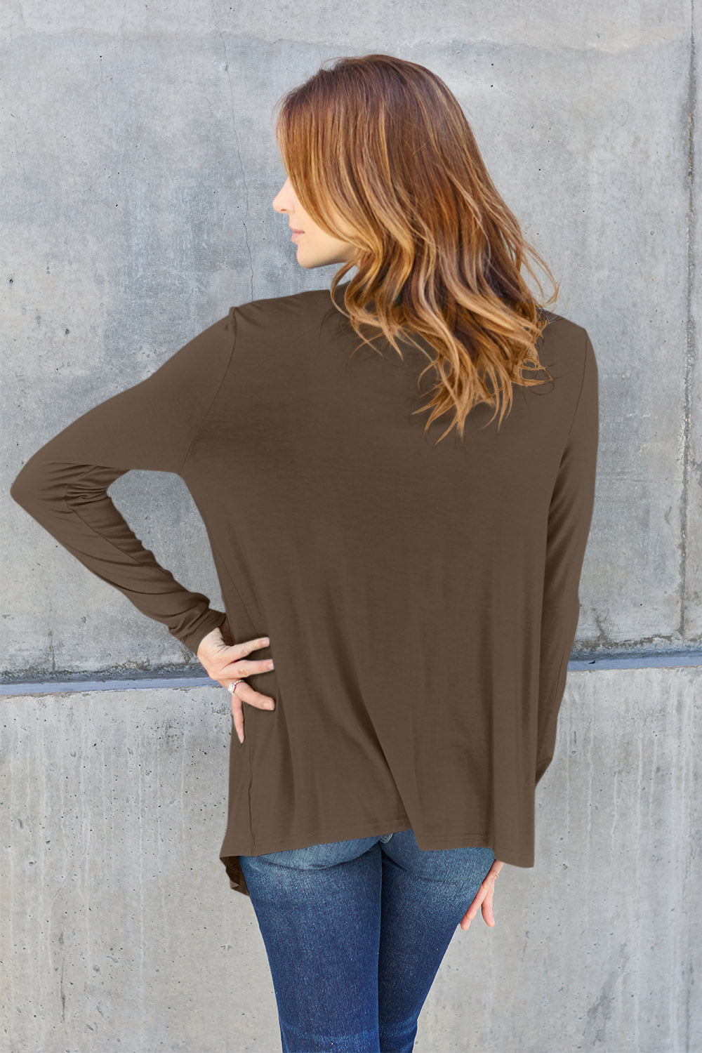 A woman wearing the Basic Bae Full Size Open Front Long Sleeve Cardigan in navy blue, paired with a white top and blue jeans, stands against a concrete wall. She touches her hair and gazes to the side, embodying a chic yet simple style. The slightly stretchy materials of the cardigan ensure comfort while maintaining its stylish simplicity. For easy care, machine wash it cold.