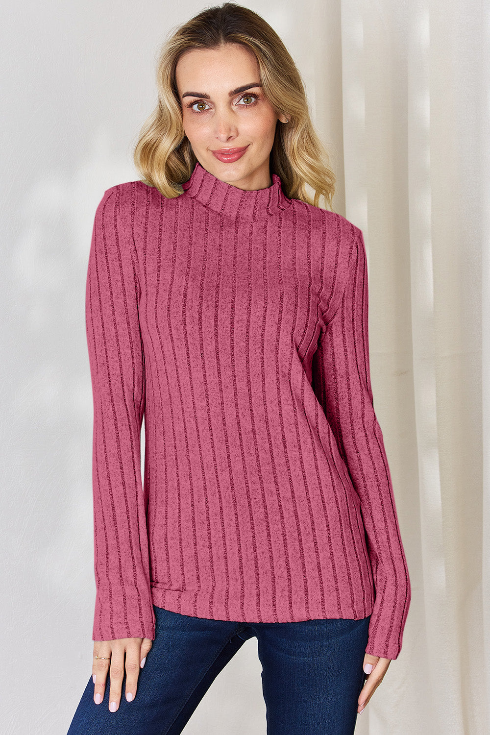 A person with long, wavy blonde hair wears a Basic Bae Full Size Ribbed Mock Neck Long Sleeve T-Shirt in pink stripes and dark jeans made of a comfortable polyester blend, standing in front of a white curtain.