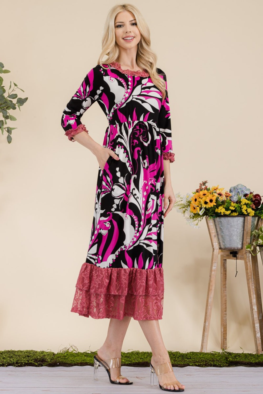 A woman wearing the Celeste Full Size Paisley Print Lace Ruffled Midi Dress poses gracefully for the camera indoors, standing beside metal planters filled with flowers.