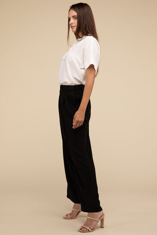 A woman stands against a beige background, highlighting her chic ensemble with a tailored fit. She wears a white short-sleeve shirt paired with versatile pink Waffle Trouser Pants, hands casually in pockets, complemented by stylish strappy sandals.