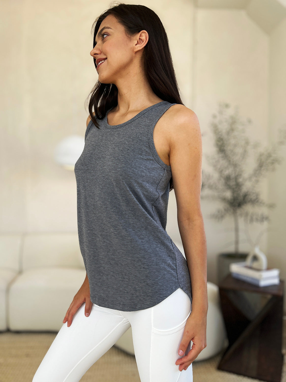 A woman wearing sunglasses, blue jeans, and the Basic Bae Full Size Round Neck Curved Hem Tank is standing in front of a concrete wall, holding her glasses with one hand and carrying a beige shoulder bag. Her basic style complements the slightly stretchy fabric of her outfit, ensuring comfort while maintaining chic simplicity.