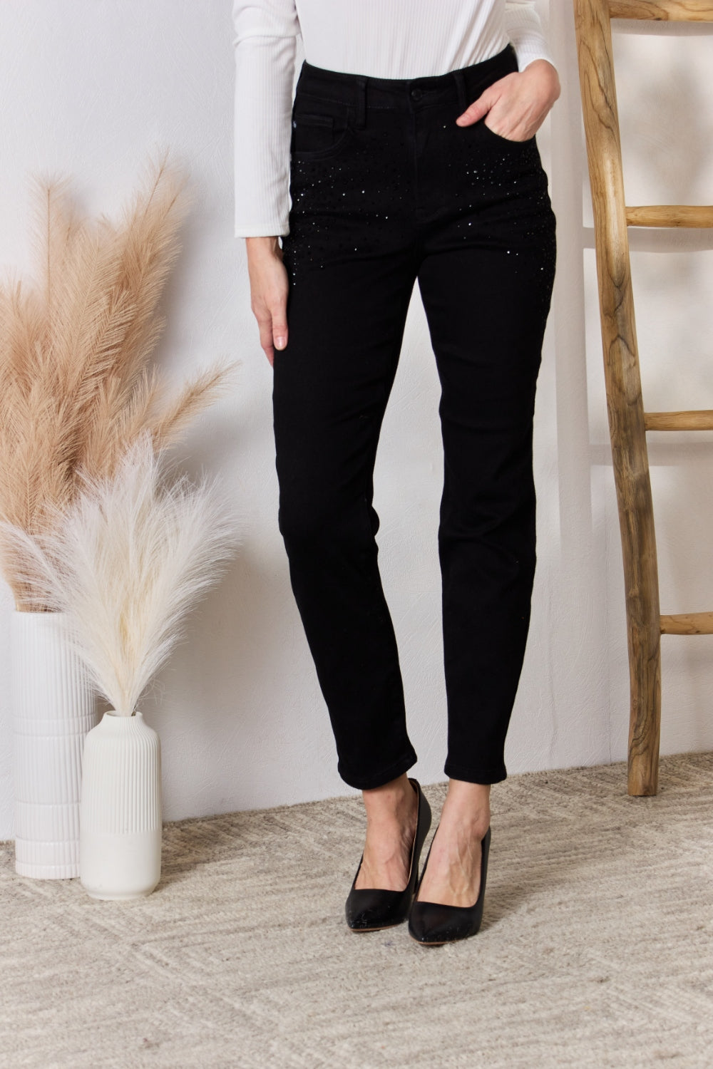 A person wearing slightly stretchy, black Judy Blue Full Size Rhinestone Embellished Slim Jeans and black shoes stands on a textured floor next to a white vase with brown and white ornamental grasses and a wooden ladder.