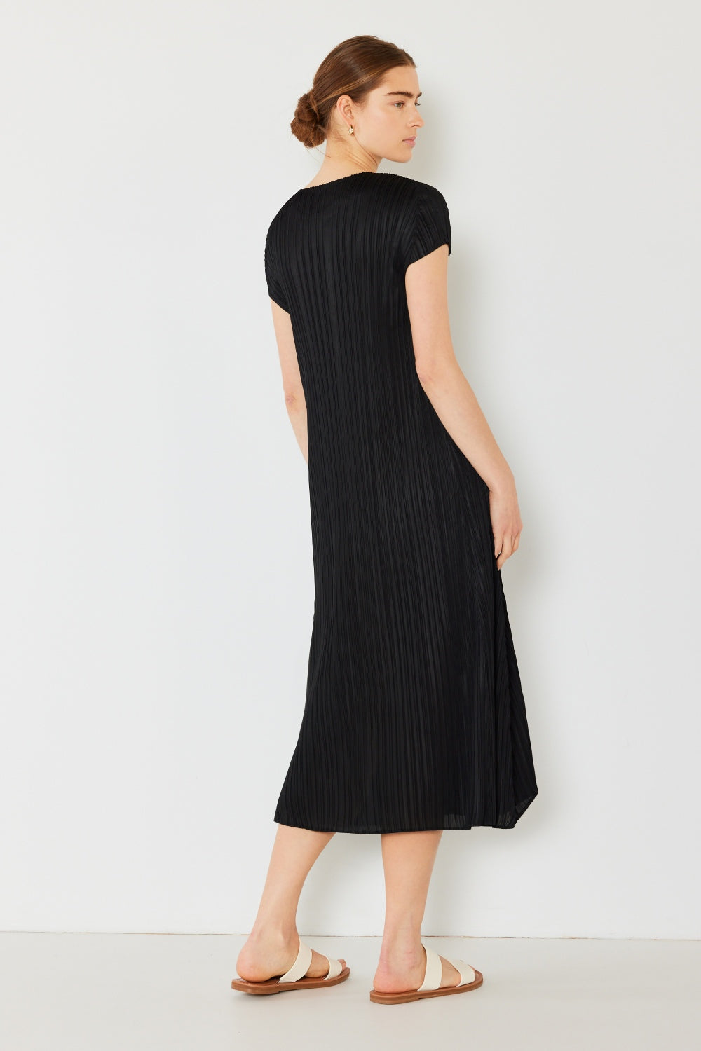 A person stands against a light wall, wearing an elegant Marina West Swim Pleated Cap Sleeve A-Line Dress in black and white sandals.