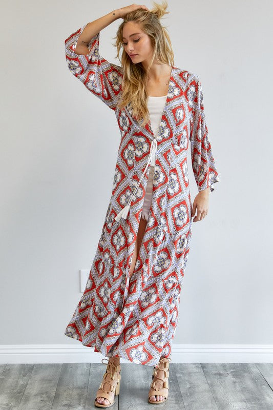 A person wearing a Printed Long Sleeve Loose Kimono with a bohemian blue and pink print over a white outfit stands in a room with a gray floor and white wall.