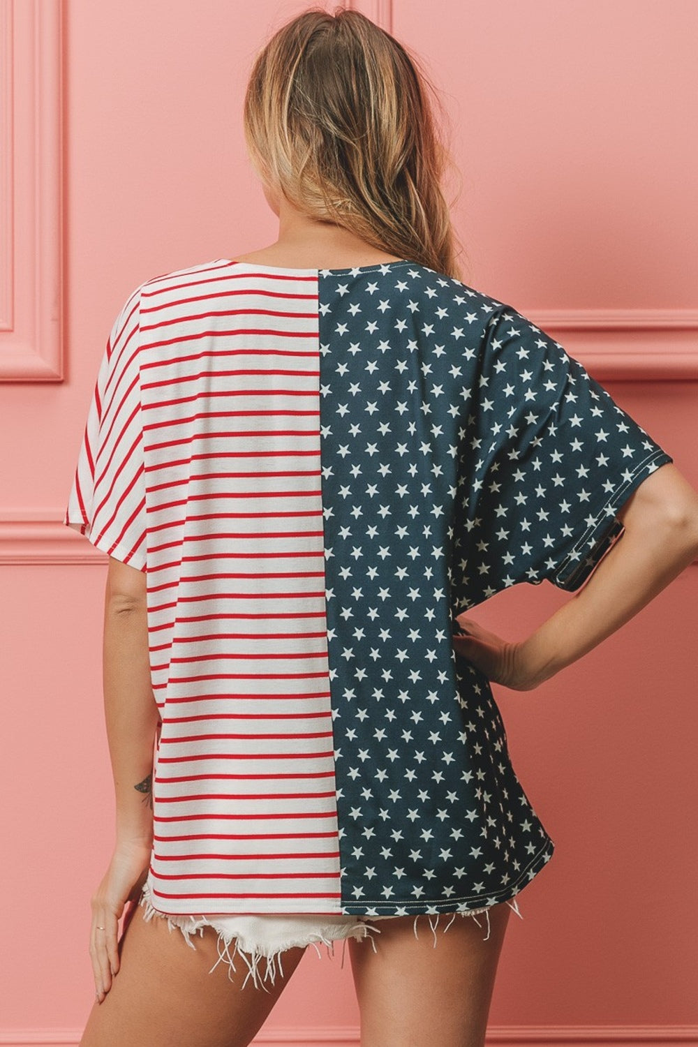 A woman stands against a pink wall, wearing the BiBi US Flag Themed Color Block Short Sleeve T-Shirt, which features one half blue with white stars and the other half red and white stripes. She is smiling with long wavy hair, showcasing her American pride casual wear.