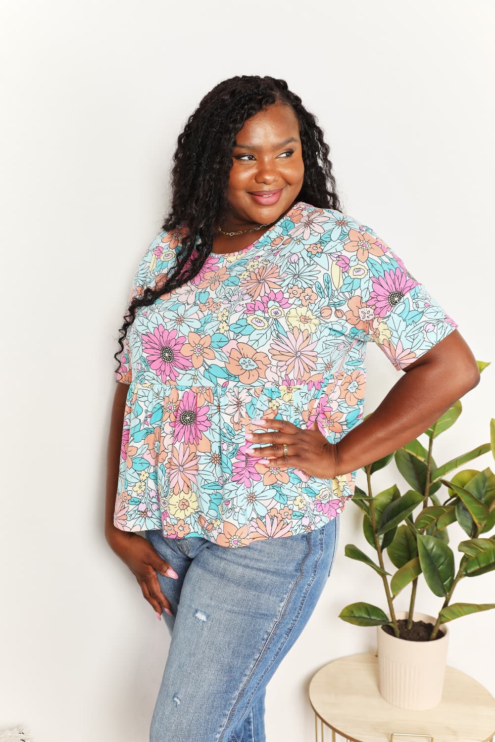 A person wearing a Double Take Floral Round Neck Babydoll Top and jeans stands with one hand in their pocket, with a plant in the background.