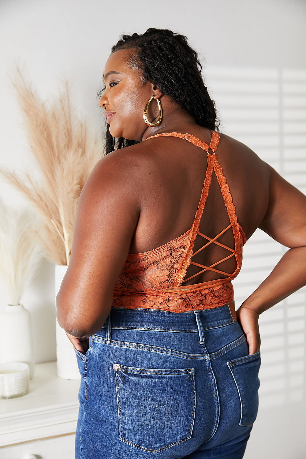 A woman with long curly hair wears the JadyK Juliette Full Size Crisscross Lace Bralette paired with blue jeans, standing beside a white vase with dried plants.