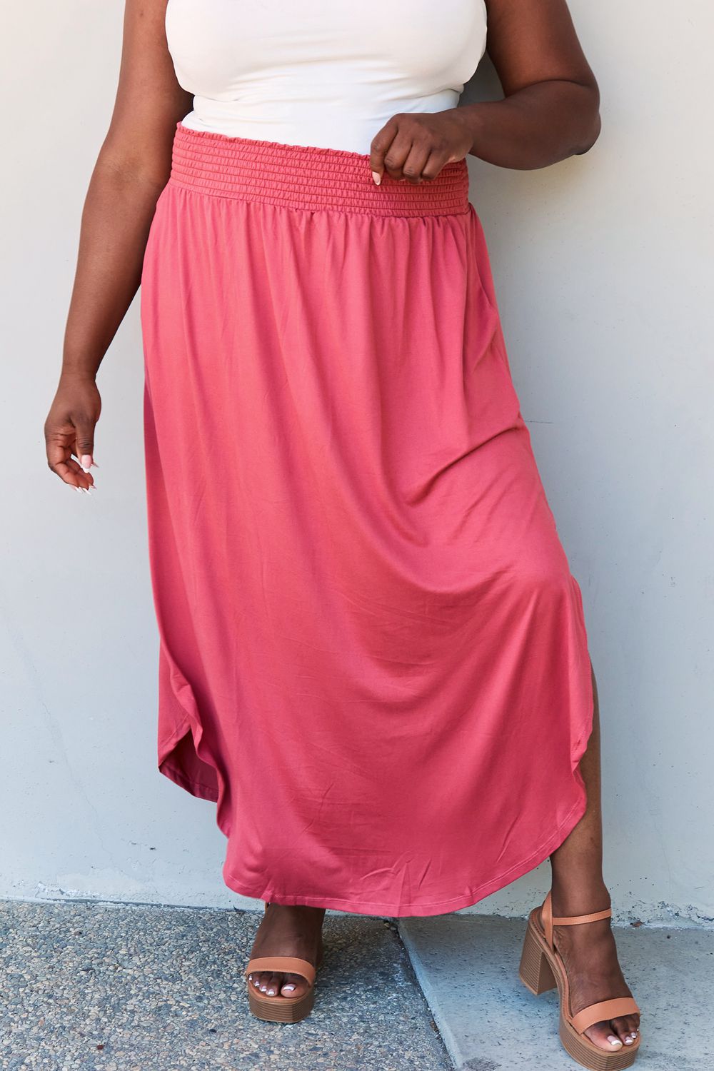 A person wearing a white top and the Doublju Comfort Princess Full Size High Waist Scoop Hem Maxi Skirt in an elegant coral color, paired with tan sandals, stands in front of a plain wall.