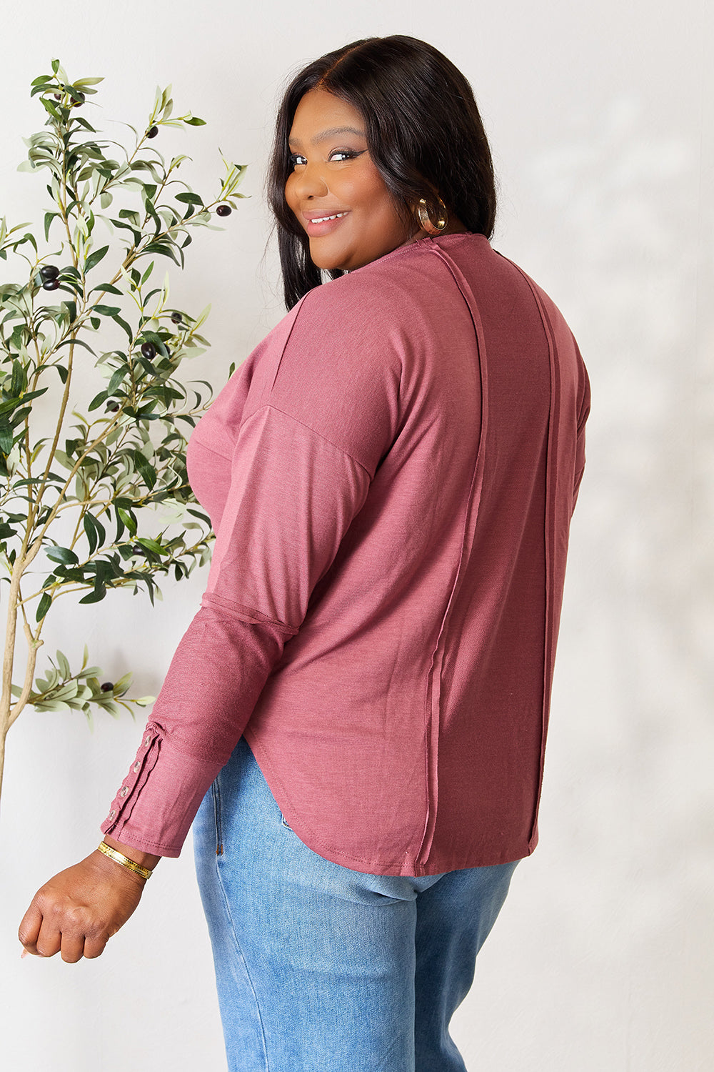 A person stands in front of a plain backdrop with a plant, wearing a maroon Culture Code Full Size V-Neck Exposed Seam Long Sleeve Blouse and blue jeans, smiling slightly with their hand on their hip. The relaxed fit of the polyester blend adds to the casual yet stylish look.