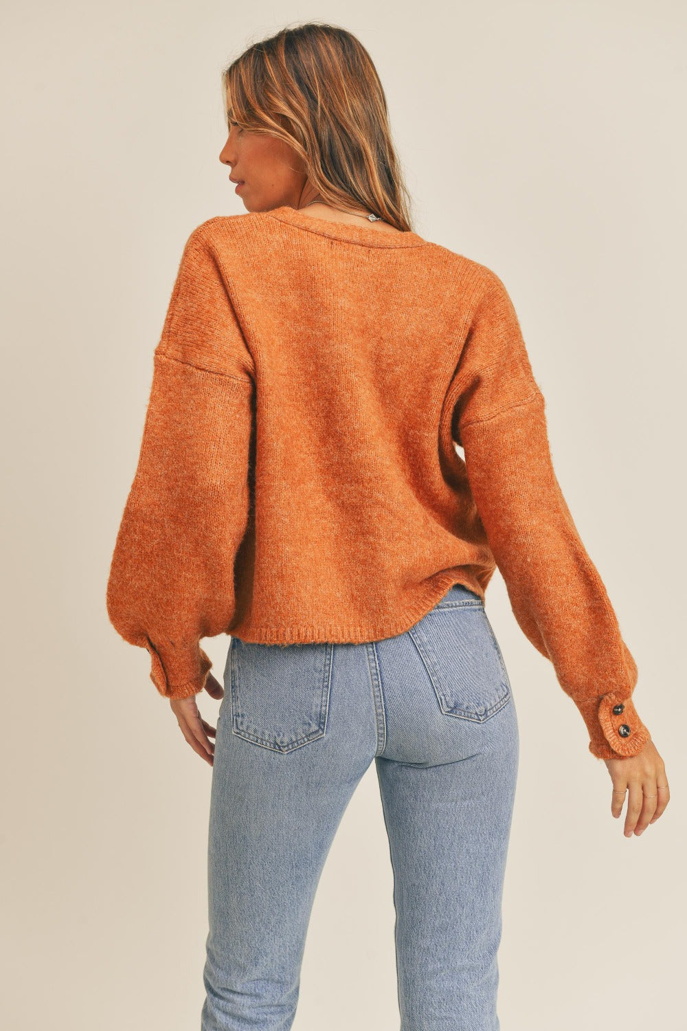 A woman wearing a MABLE Long Sleeve Button Down Sweater Cardigan in burnt orange and light blue jeans looks to her left against a plain background, exuding classic elegance.