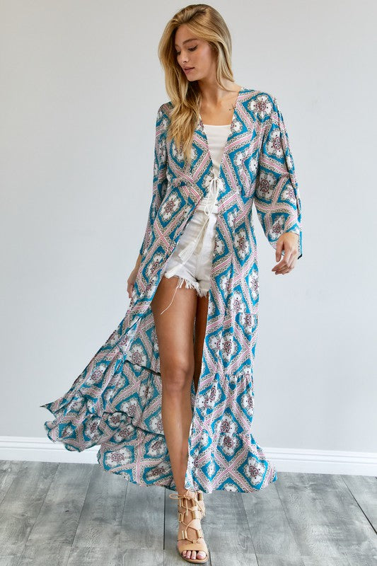 A person wearing a Printed Long Sleeve Loose Kimono with a bohemian blue and pink print over a white outfit stands in a room with a gray floor and white wall.