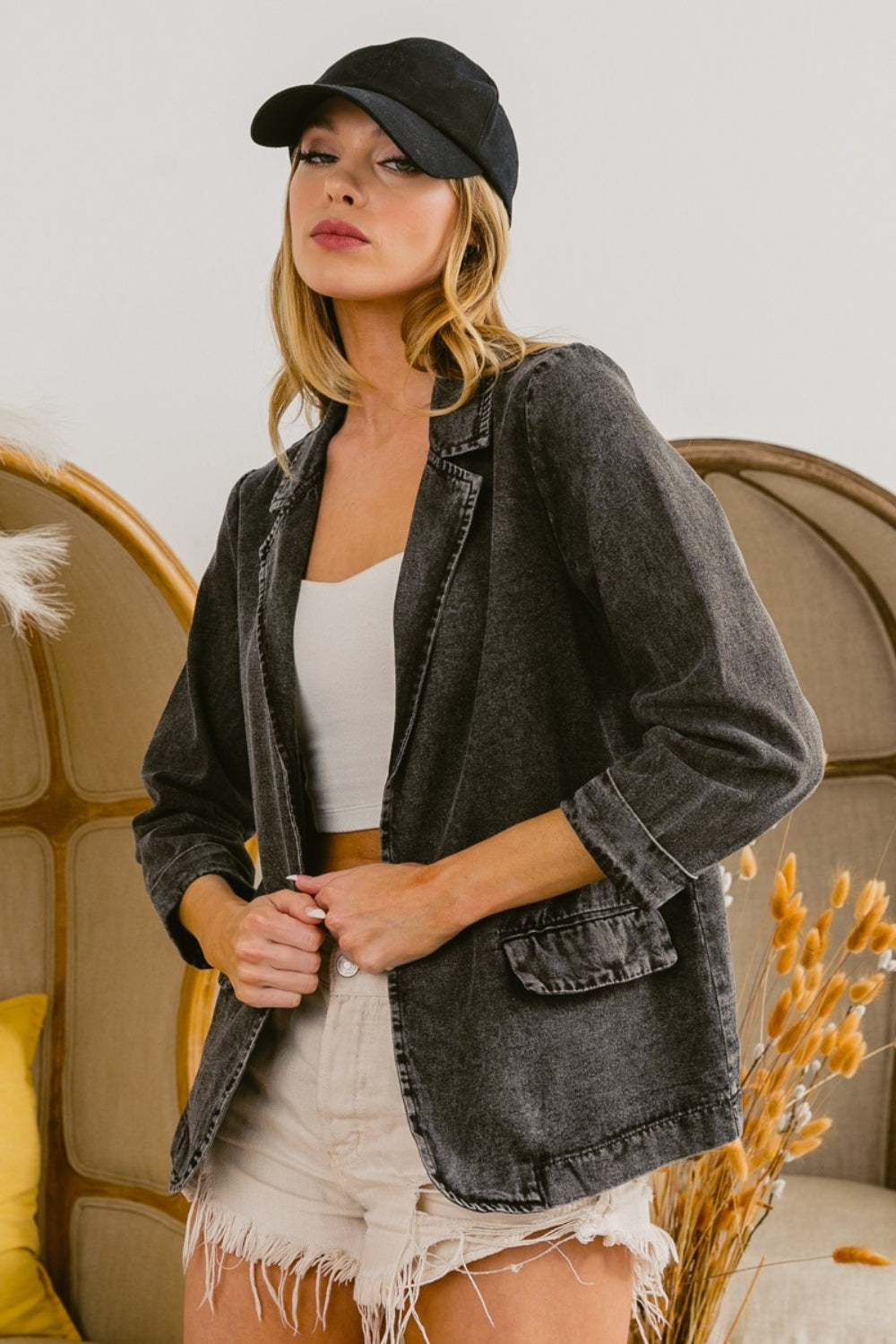 A woman stands indoors wearing a versatile, stylish wardrobe piece: the BiBi Single Breasted Washed Denim Blazer in black, paired with a white top, khaki shorts, and a black cap. She has her hands in her blazer pockets and looks slightly off to the side.