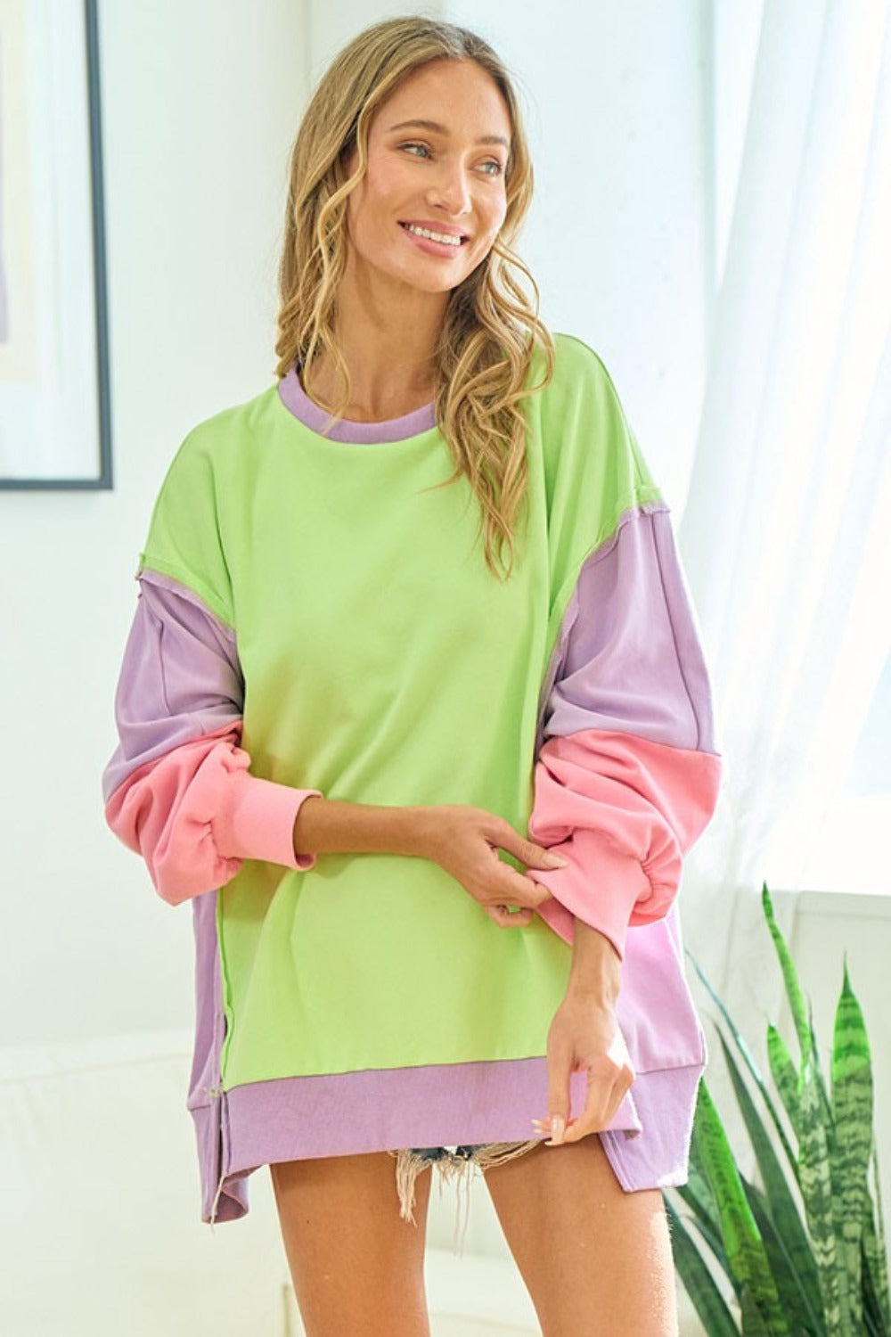 A woman wearing the First Love Color Block Long Sleeve Slit Sweatshirt and matching shorts poses indoors, smiling with one hand touching her hair.