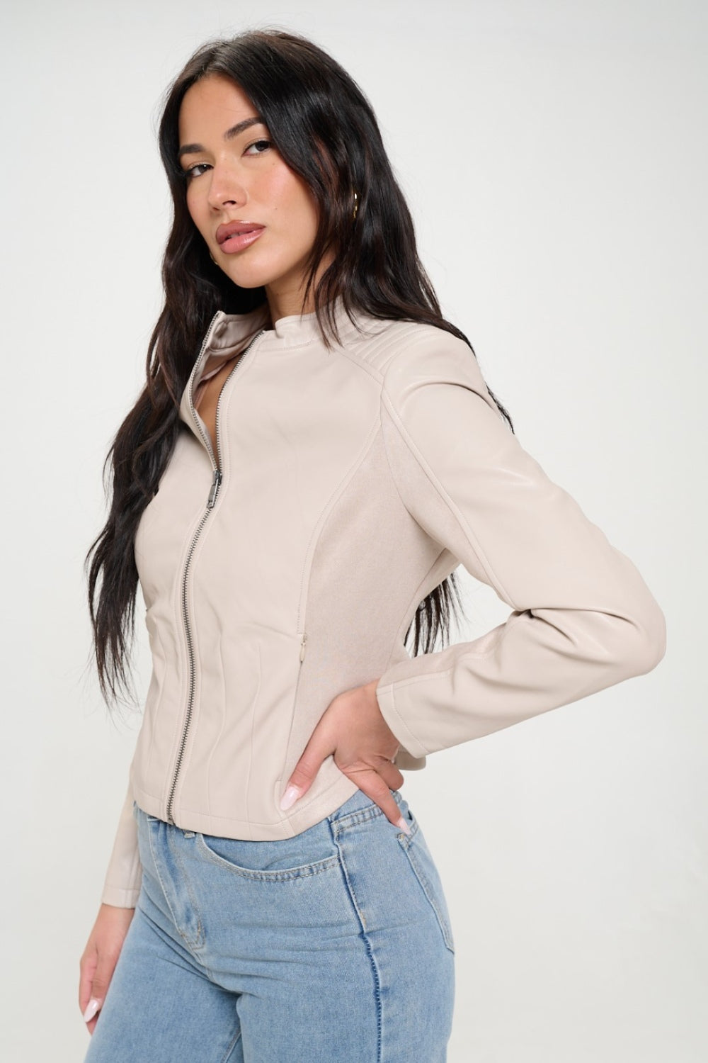 A woman with long dark hair, wearing the cruelty-free Coalition LA Zip Up Vegan Moto Jacket in light beige, pairs it with blue jeans as she stands with her arms crossed.