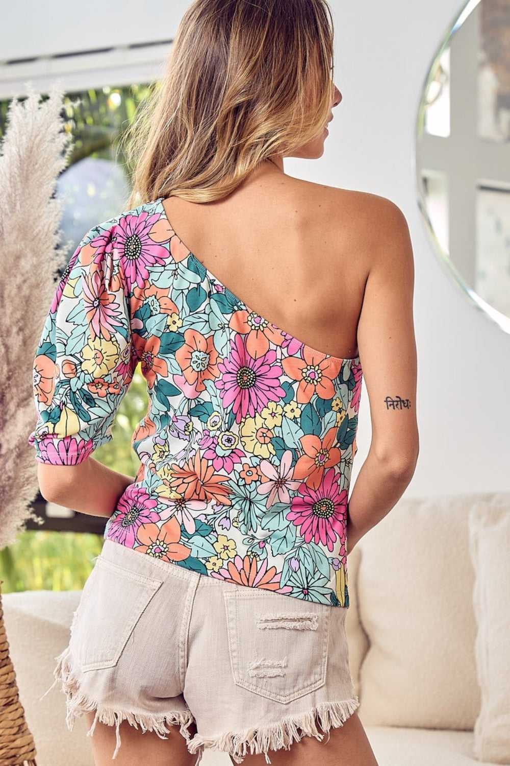 A smiling woman in a BiBi Floral Puff Sleeve One Shoulder Top, paired with beige shorts, stands indoors near a window.