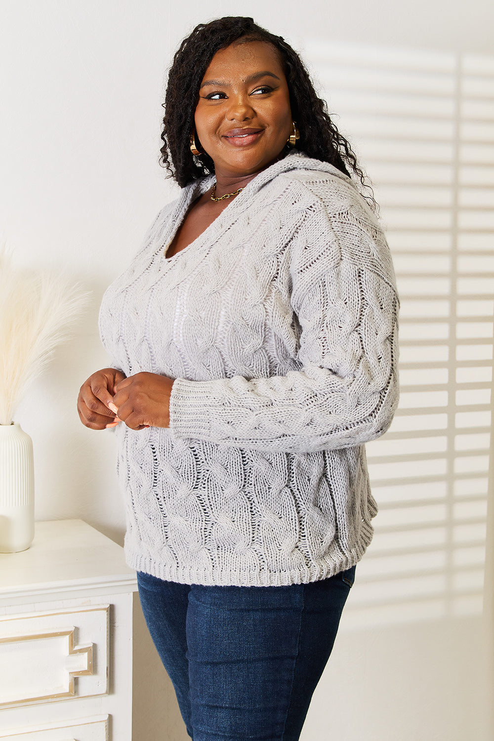 A person with long hair wearing a cozy Woven Right Cable-Knit Hooded Sweater and dark jeans is standing indoors looking to the side.