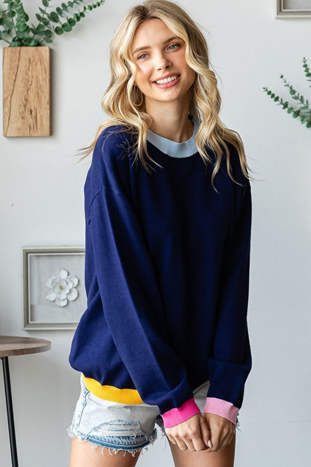 A person with long blonde hair wearing a First Love Contrast Ribbed Round Neck Long Sleeve Sweater and light denim shorts stands indoors near a small table and plant.