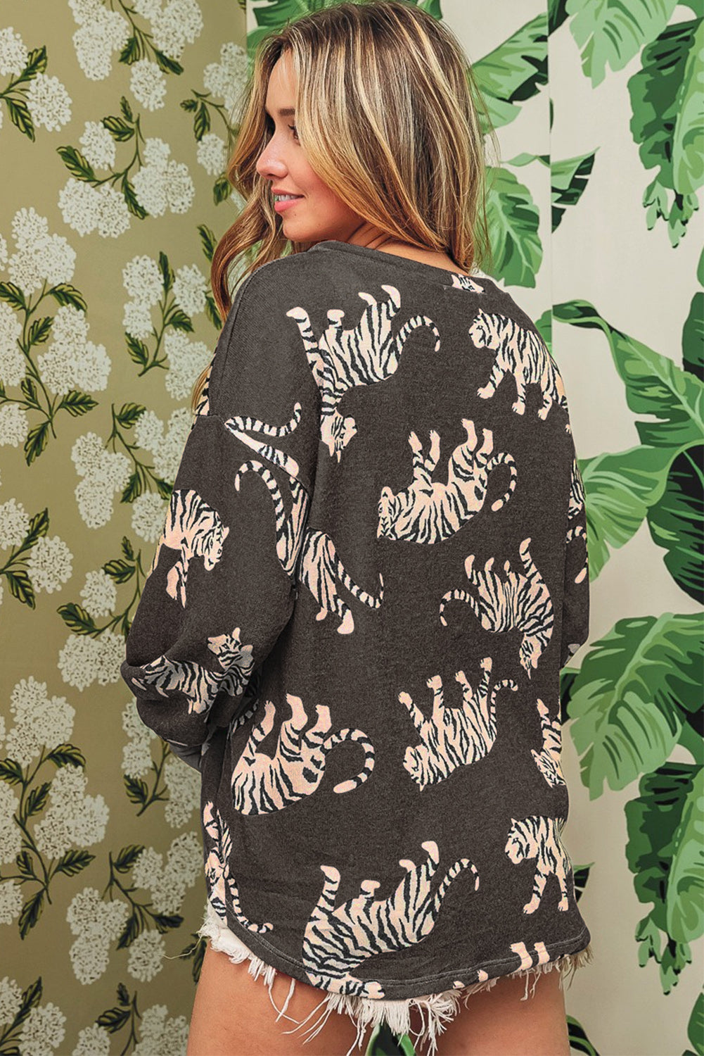 A person with long hair, wearing the Lively Tiger Print Casual Sweatshirt, stands in front of tropical-themed wallpaper. The backdrop features green leaves and white flowers, perfectly complementing the unique sweatshirt adorned with a striking white tiger pattern.