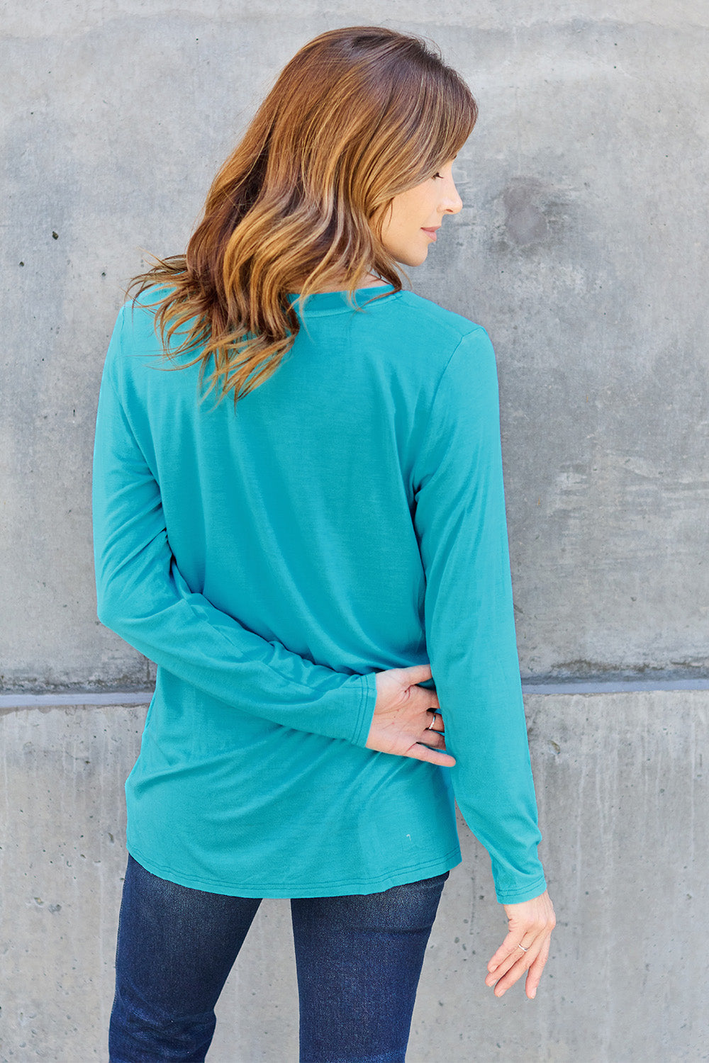 A woman with light brown hair stands against a concrete wall, wearing the Basic Bae Full Size V-Neck Long Sleeve Top in olive green and blue jeans, with a white backpack on her shoulder. Her outfit exudes basic style and is made from polyester, ensuring it’s both comfortable and suitable for machine wash cold. She is looking at the camera.