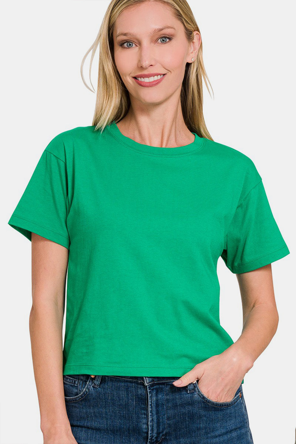 A person wearing a Zenana Round Neck Short Sleeve Cropped T-Shirt in green and blue jeans is smiling with a hand in the pocket against a white background.