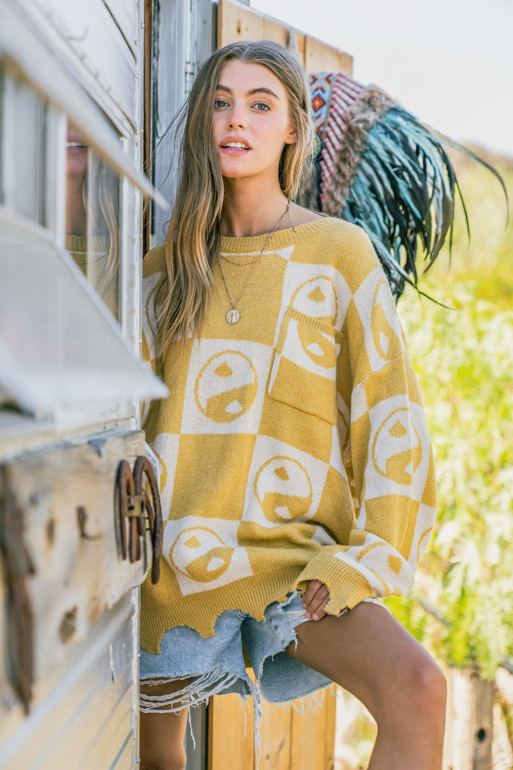 A woman wearing the And The Why Checkerboard Sweater with a Yin Yang pattern and distressed denim shorts stands in a doorway.