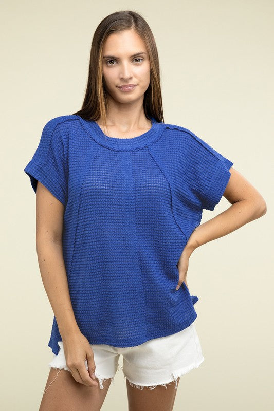 A woman with long brown hair is wearing a purple Brushed Waffle Exposed-Seam Short Sleeve Top and blue jeans, standing against a plain beige background. This casual wardrobe choice, with its short sleeves and side slits, is perfect for everyday wear.